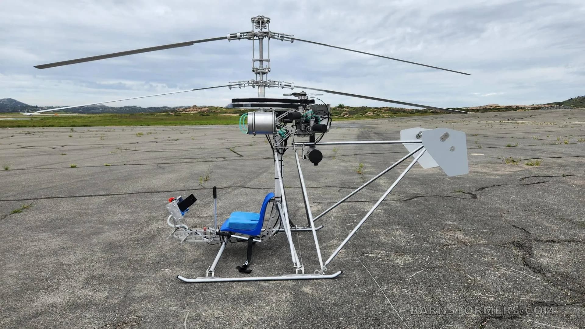 A single-seat ultralight helicopter with a blue seat is parked on a cracked asphalt surface under a cloudy sky.