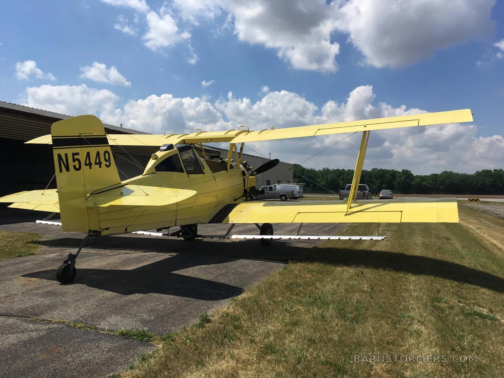 A yellow biplane crop duster, tail number N5449, viewed from the rear-right, parked on an airfield with distant vehicles.
