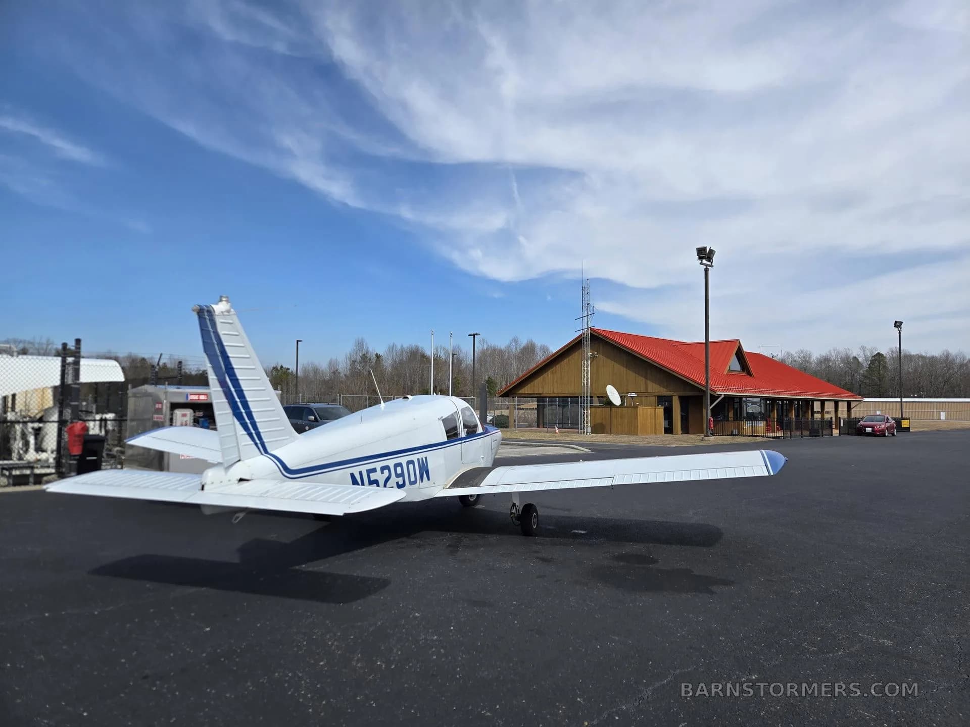 A white and blue single-engine aircraft (N5290W) parked on an asphalt ramp with a red-roofed building.