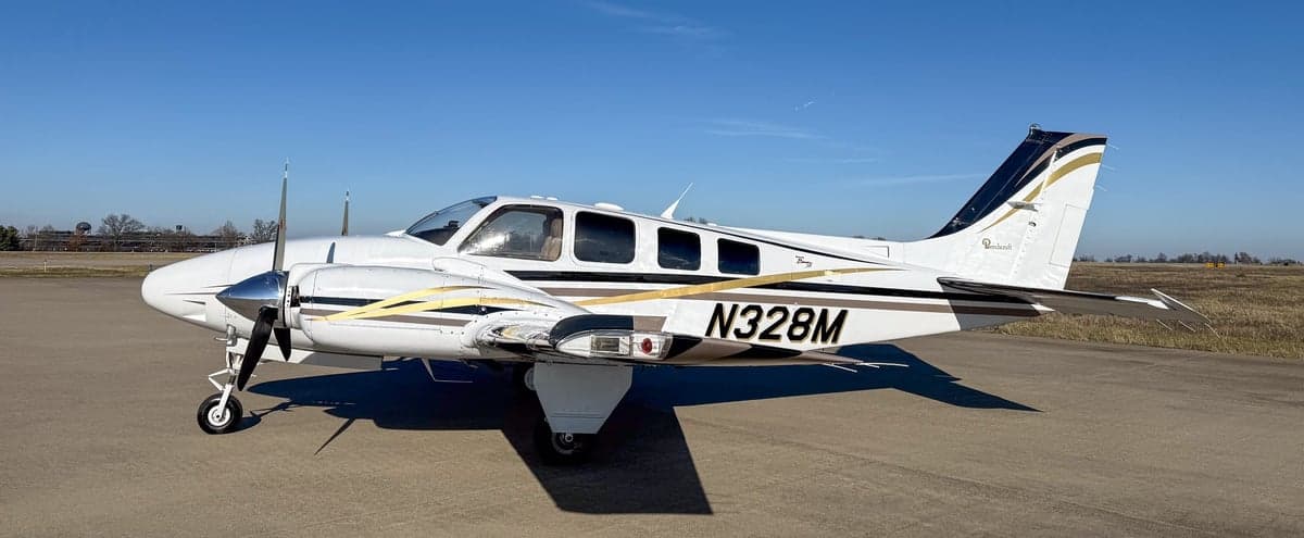 Side view of a white twin-engine propeller aircraft N328M with black and gold stripes on an airfield.