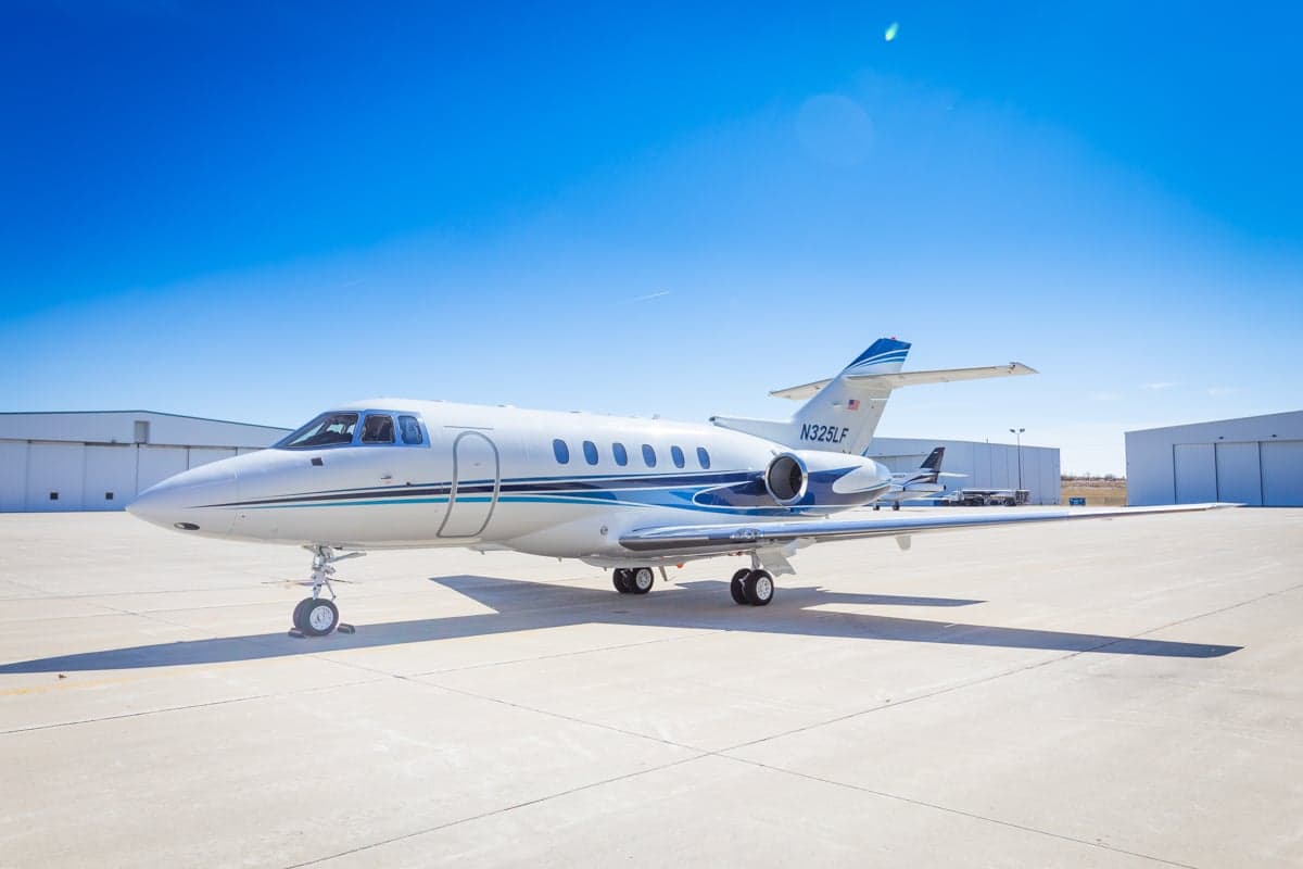 A white and blue jet sits on the tarmac in front of a hangar on a sunny day.