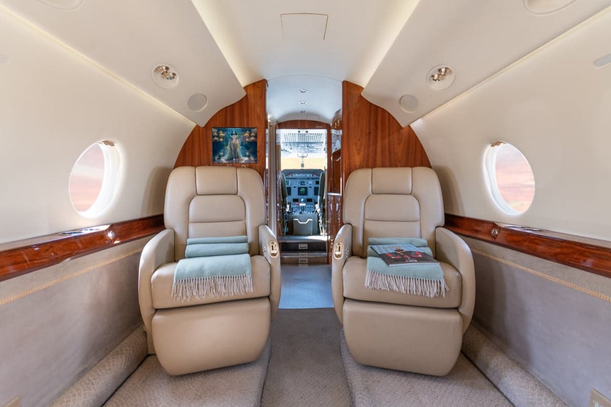 A view down the aircraft cabin aisle, showing two beige leather seats in the foreground and the cockpit entrance in the distance.