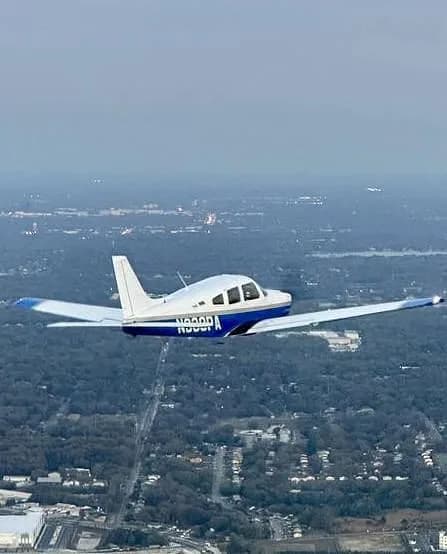 A white and blue Piper Arrow aircraft with registration N338PA flying over a city landscape with trees and buildings.