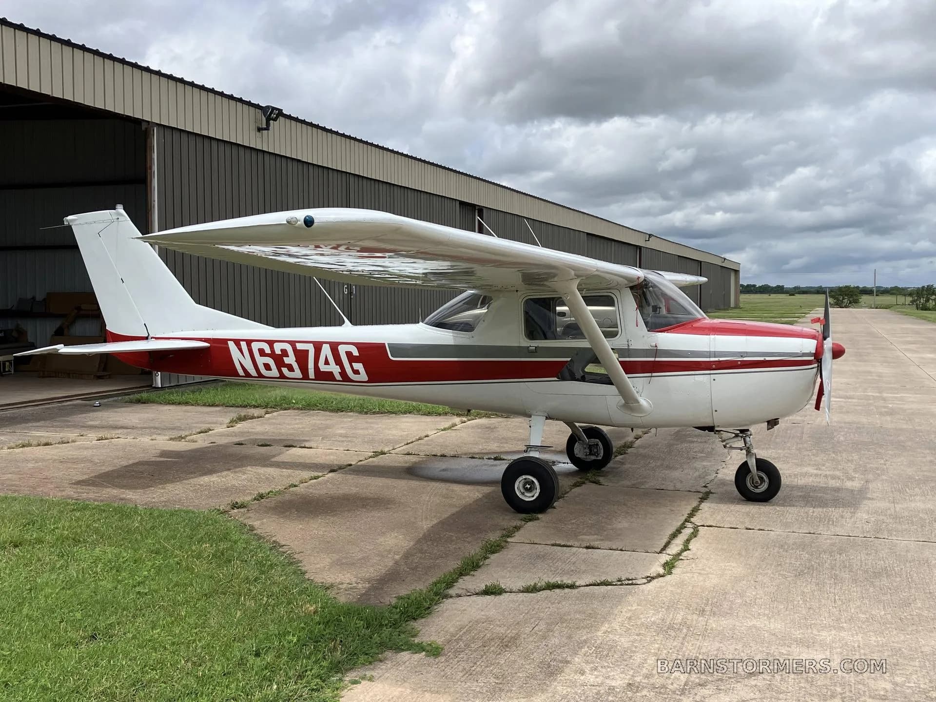 A white and red Cessna 172 aircraft with registration N6374G parked on a concrete pad in front of a hangar under a cloudy sky.