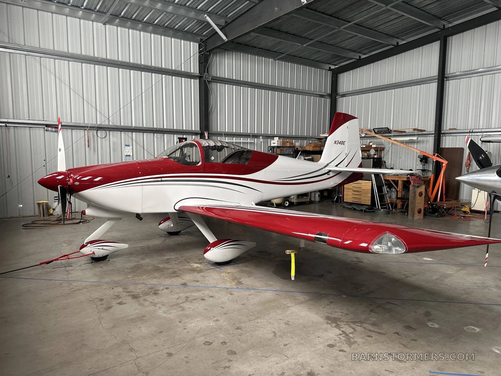 A red and white single-engine aircraft (N34BE) is parked inside a metal hangar.