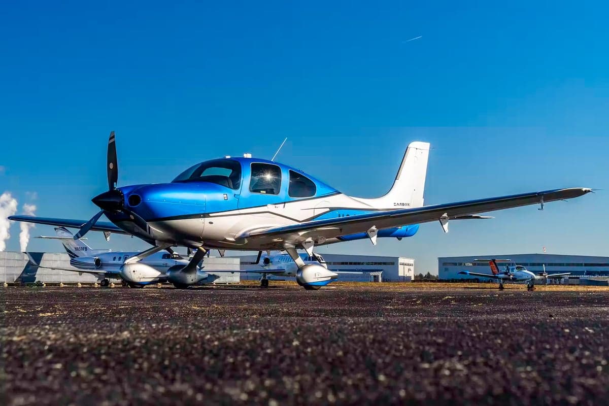 A blue and white Cirrus SR22T parked on the tarmac with other aircraft in the background.