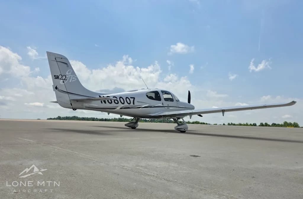 A silver Cirrus SR22 aircraft (N68007) with its doors closed, parked on an airfield, viewed from the rear-right.