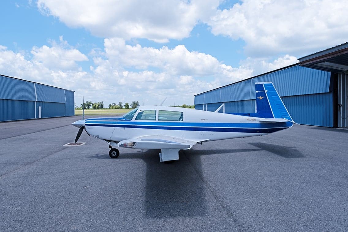 Side view of a white and blue Mooney M20F aircraft parked on the tarmac in front of a hangar.