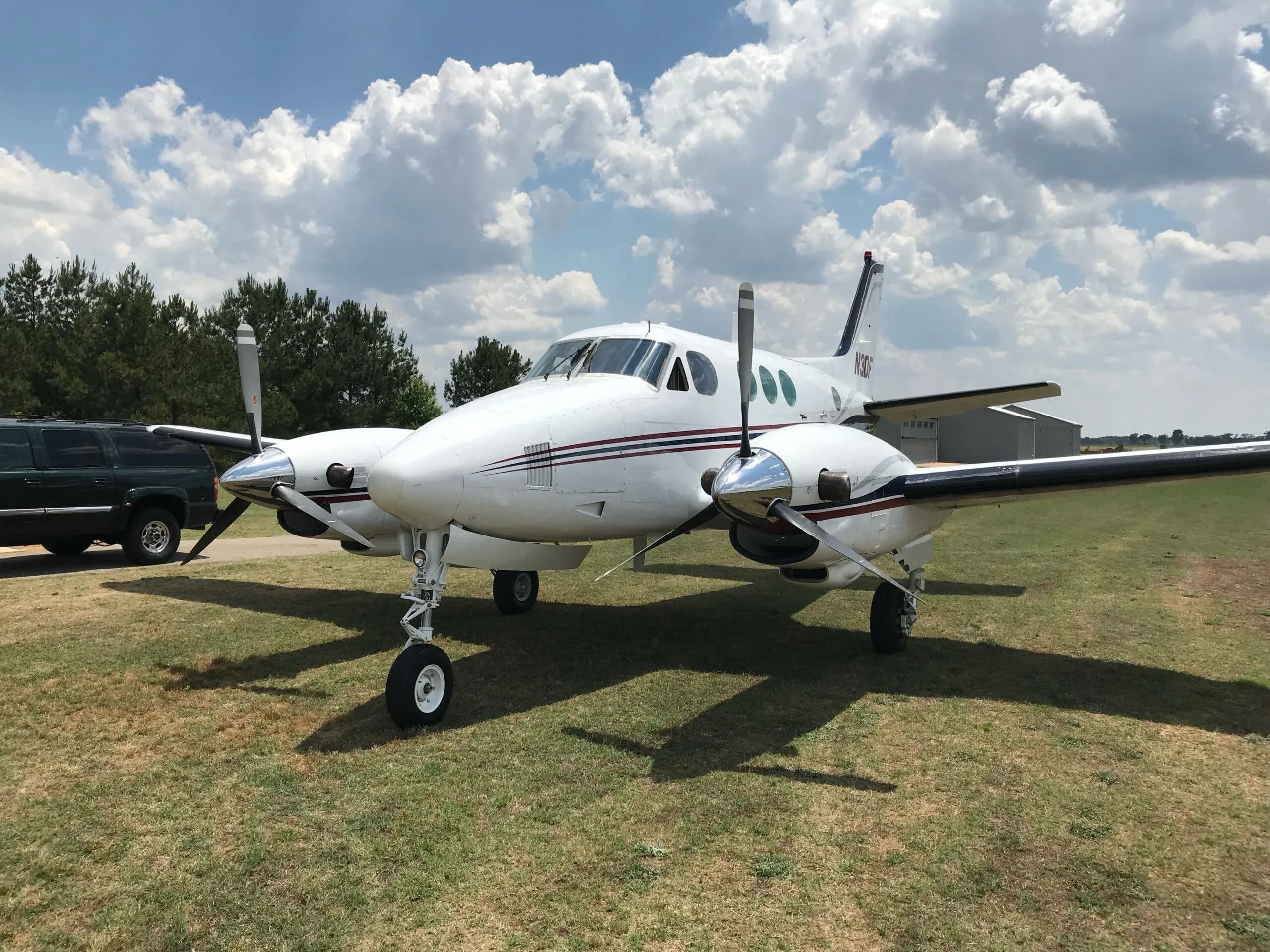 A white twin-engine turboprop aircraft, N3DF, with red and blue stripes, parked on a grassy field under a partly cloudy sky, viewed from the front-left.
