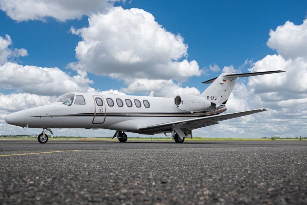 A white Cessna Citation CJ3 (D-IALI) private jet with black and gold stripes parked on an asphalt runway under a blue sky with white clouds.