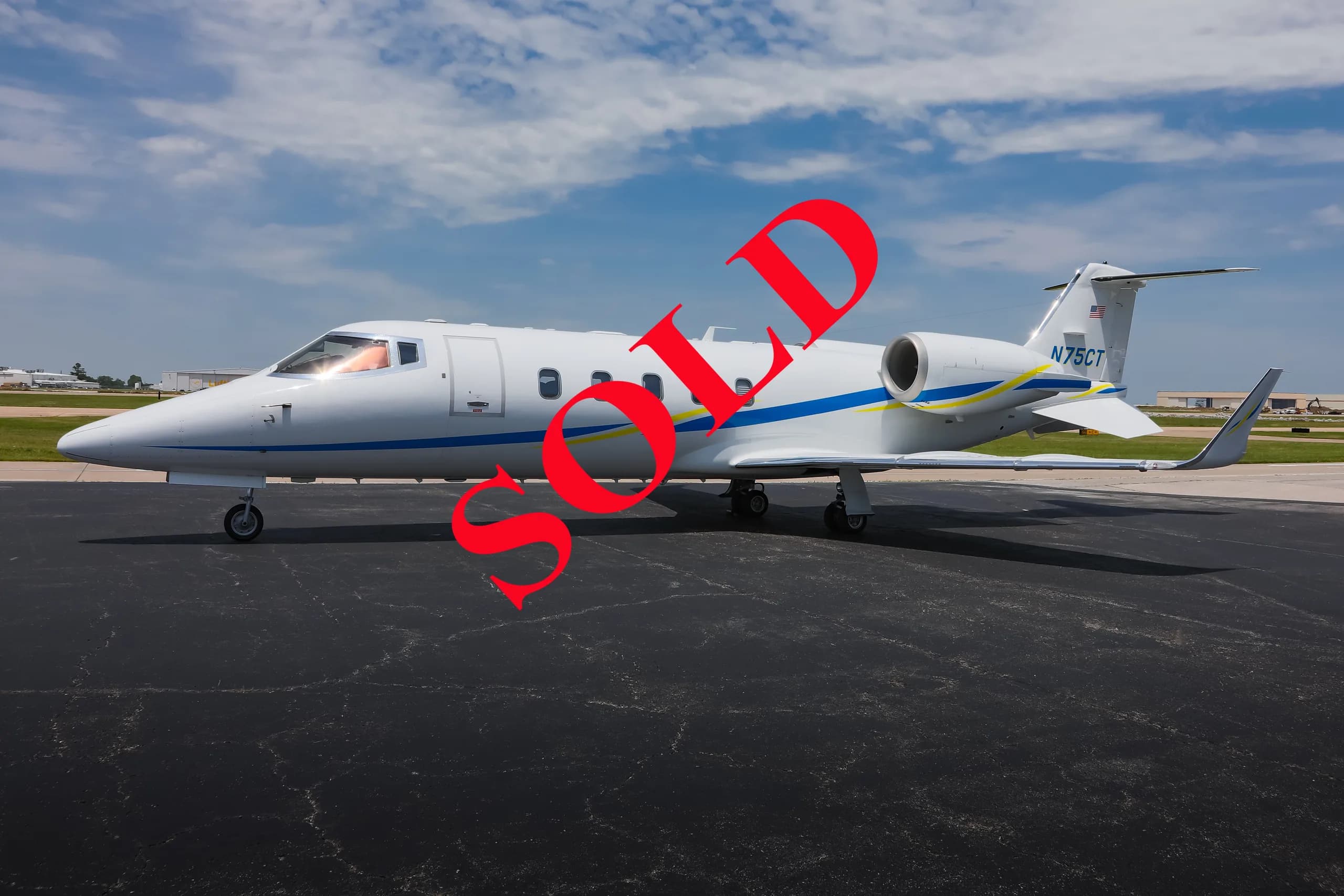 A white business jet with blue and yellow stripes, registration N75CT, parked on an asphalt tarmac under a partly cloudy sky.