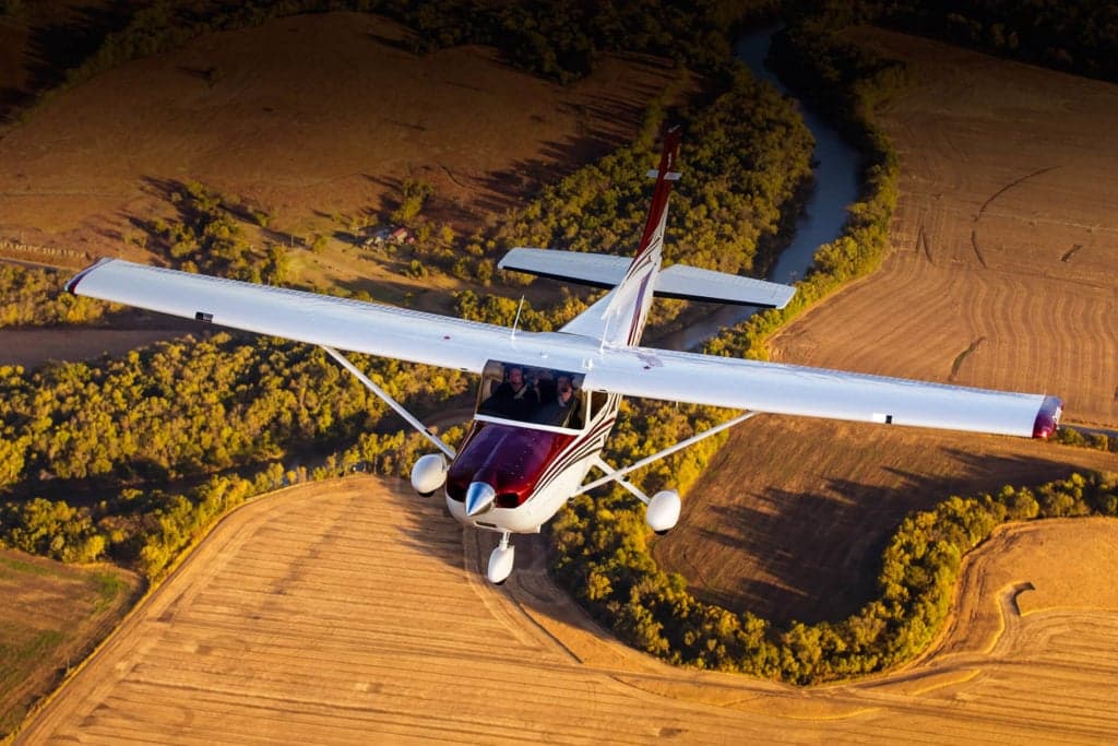 A white and maroon Cessna 182 aircraft flying over golden fields and a winding river.