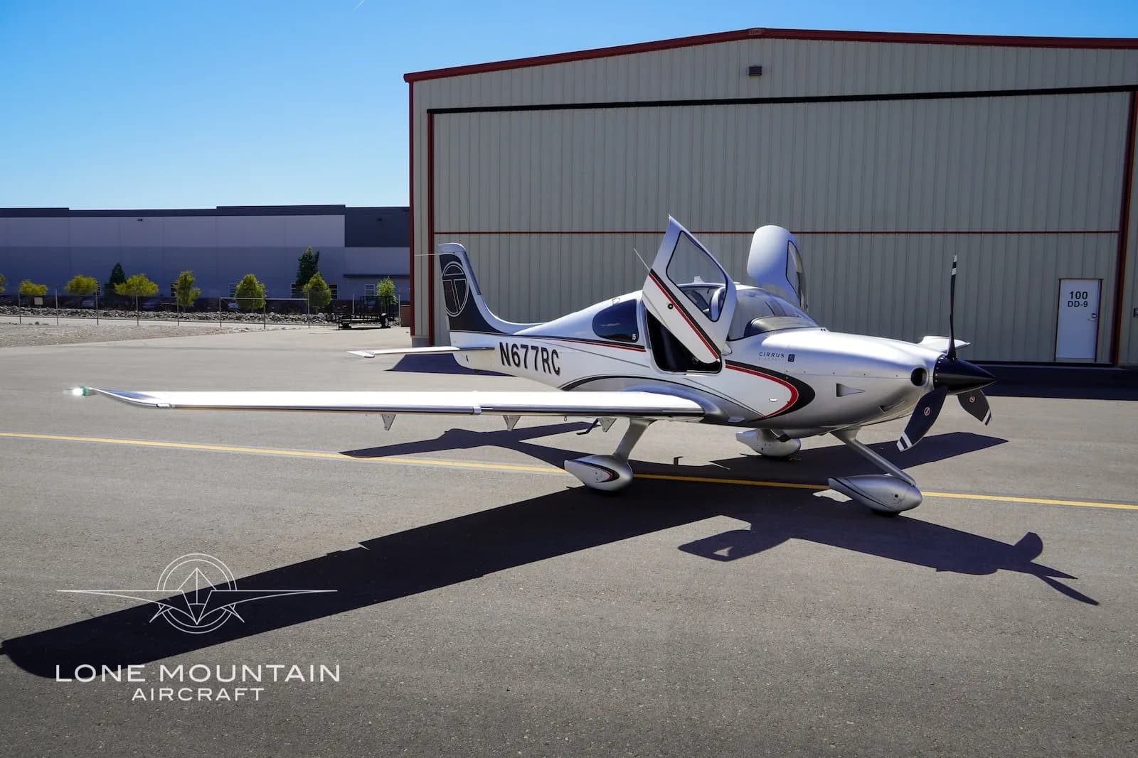 A silver Cirrus SR22 aircraft, registration N677RC, with its left door open, parked on an airfield next to a hangar.