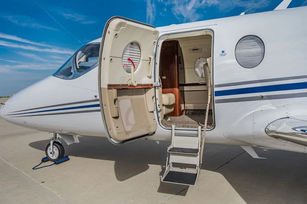 The front section of a white business jet with its main entry door open and stairs extended, revealing a glimpse of the interior.