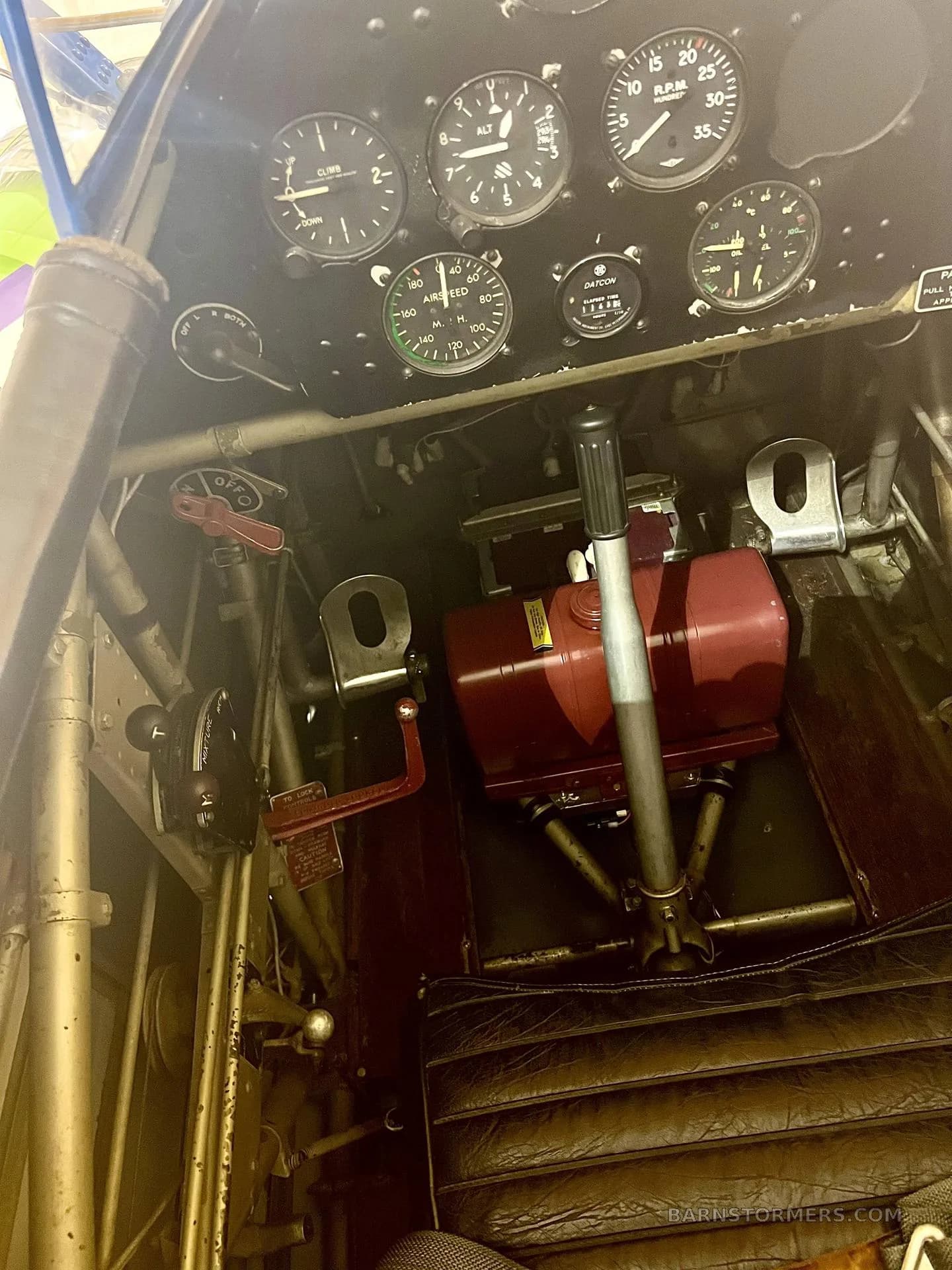 The cockpit of a biplane shows the instrument panel, controls, and a brown leather seat.