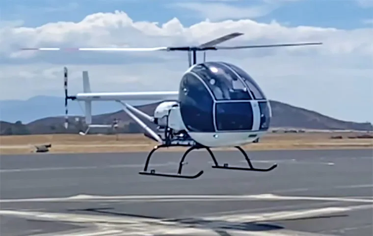 A white and black helicopter hovering above a landing pad on a sunny day with hills in the background.