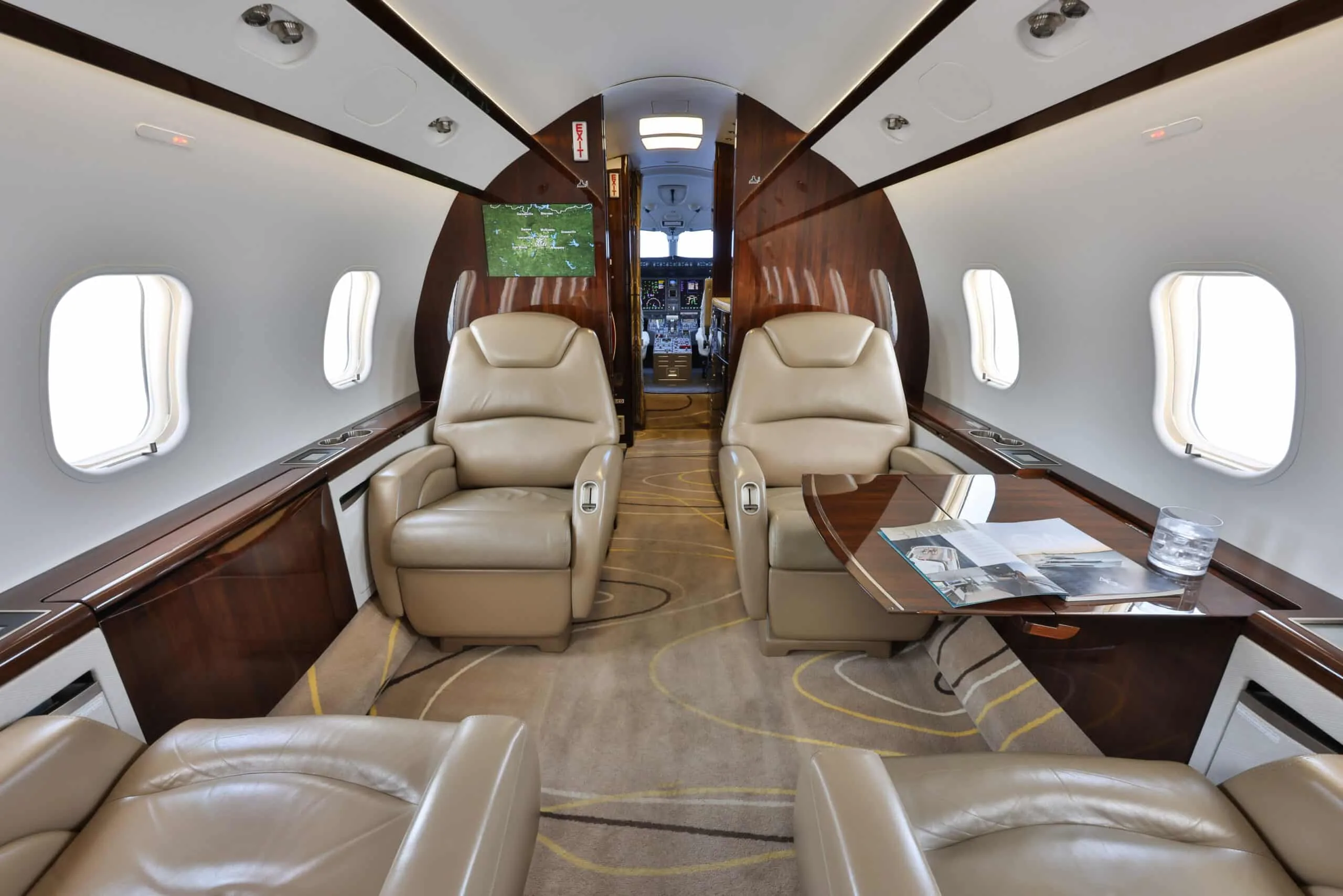 The luxurious cabin interior of a private jet features beige leather seats, dark wood paneling, and a view towards the cockpit.