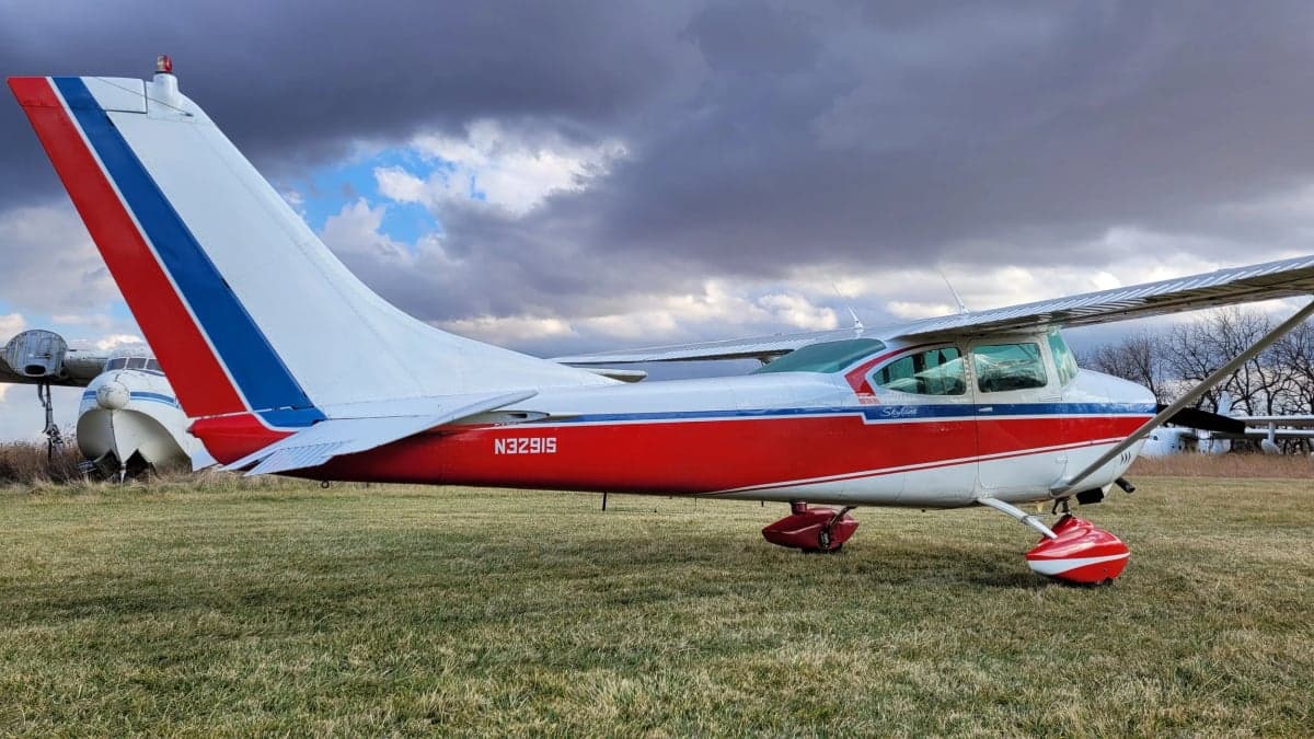 A side view of the Cessna Skylane N3291S with red, white, and blue paint scheme on a grassy field with other aircraft in the background.