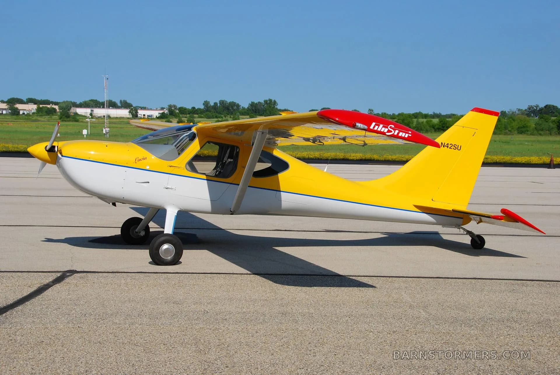 A yellow and white light sport aircraft with red wingtips and tail, registration N42SU, parked on an asphalt runway under a clear blue sky.