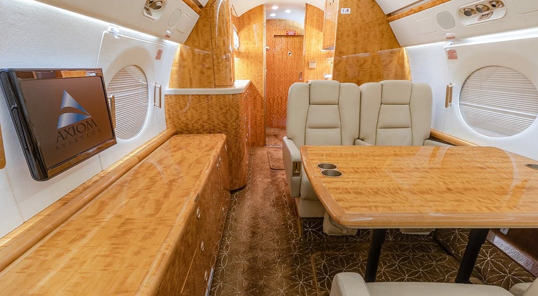 The aircraft's interior features a table, cream-colored leather seats, and a monitor displaying the Axiom Aviation logo.
