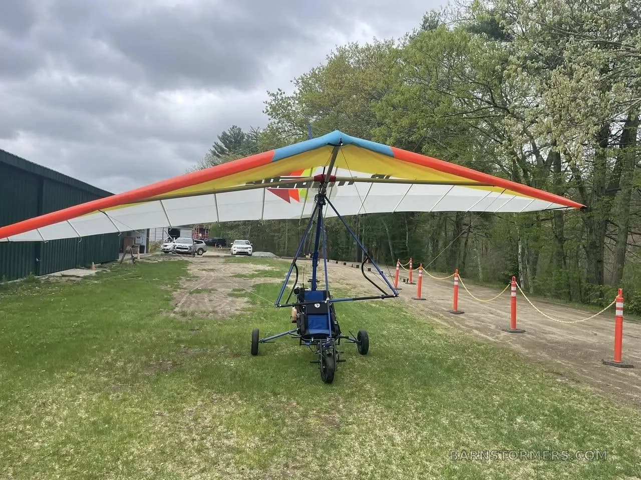 A powered hang glider with an orange, yellow, and white wing is parked on a grassy field with trees and a building in the background.