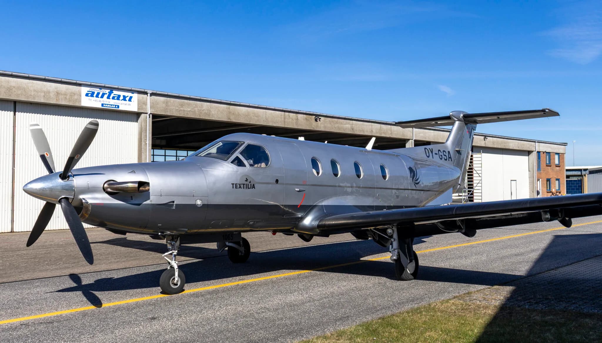 A silver Pilatus PC-12 aircraft with registration OY-GSA parked on a tarmac in front of a hangar.