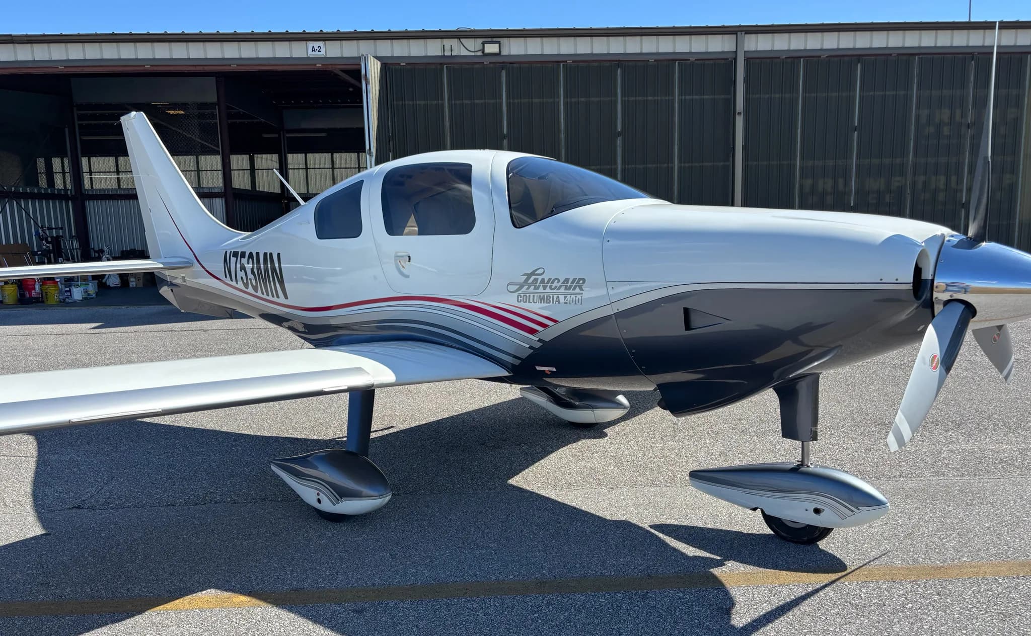Front-quarter view of the Lancair Columbia 400 N753MN with its white, grey, and red livery in front of a hangar.