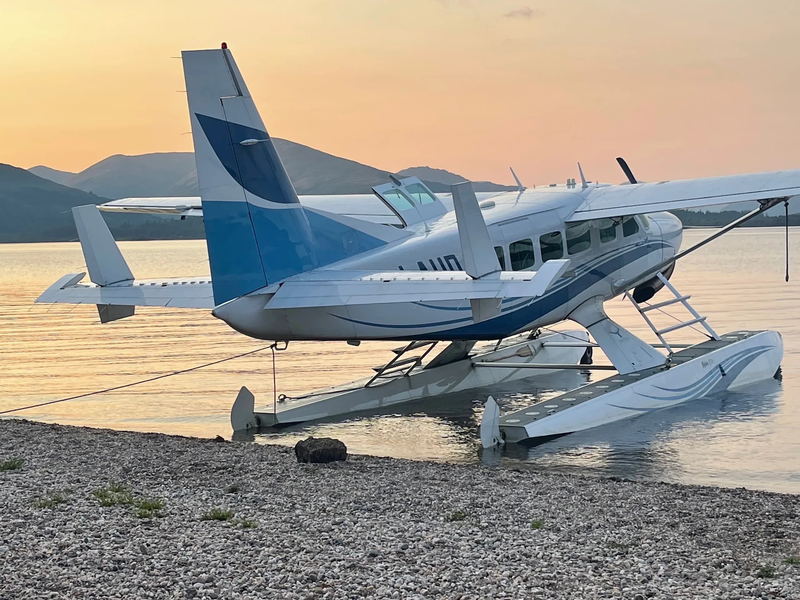 A white and blue floatplane, a Cessna Caravan, is tied to a rocky shore at sunset, with mountains in the distance.