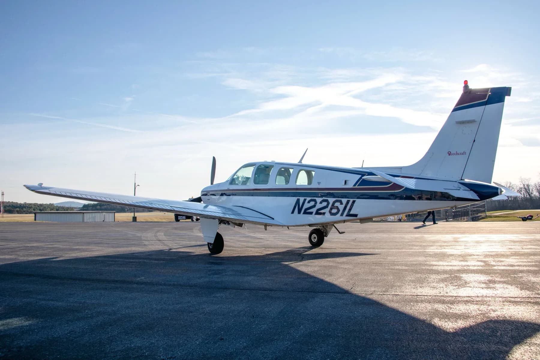 A white Beechcraft Bonanza N2261L with blue and dark blue stripes parked on an airfield, viewed from the side.