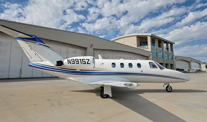 A white and blue Cessna Citation CJ1 aircraft with registration N9915Z parked on the tarmac in front of a hangar on a sunny day.