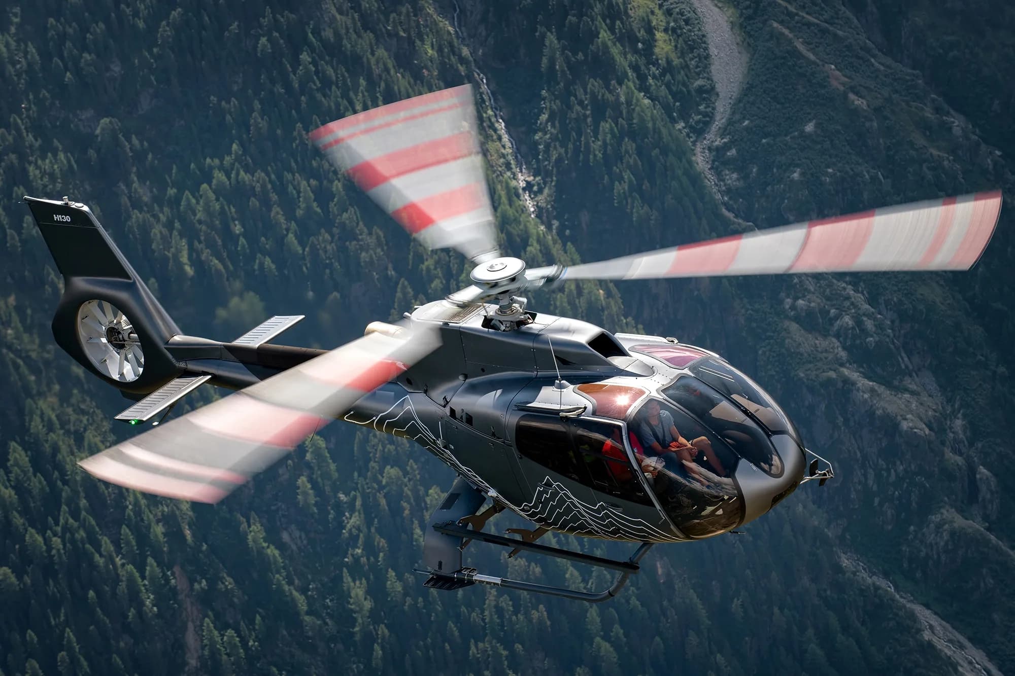 A dark grey H130 helicopter with white mountain graphics and red-striped rotor blades flies over a forested mountain range.