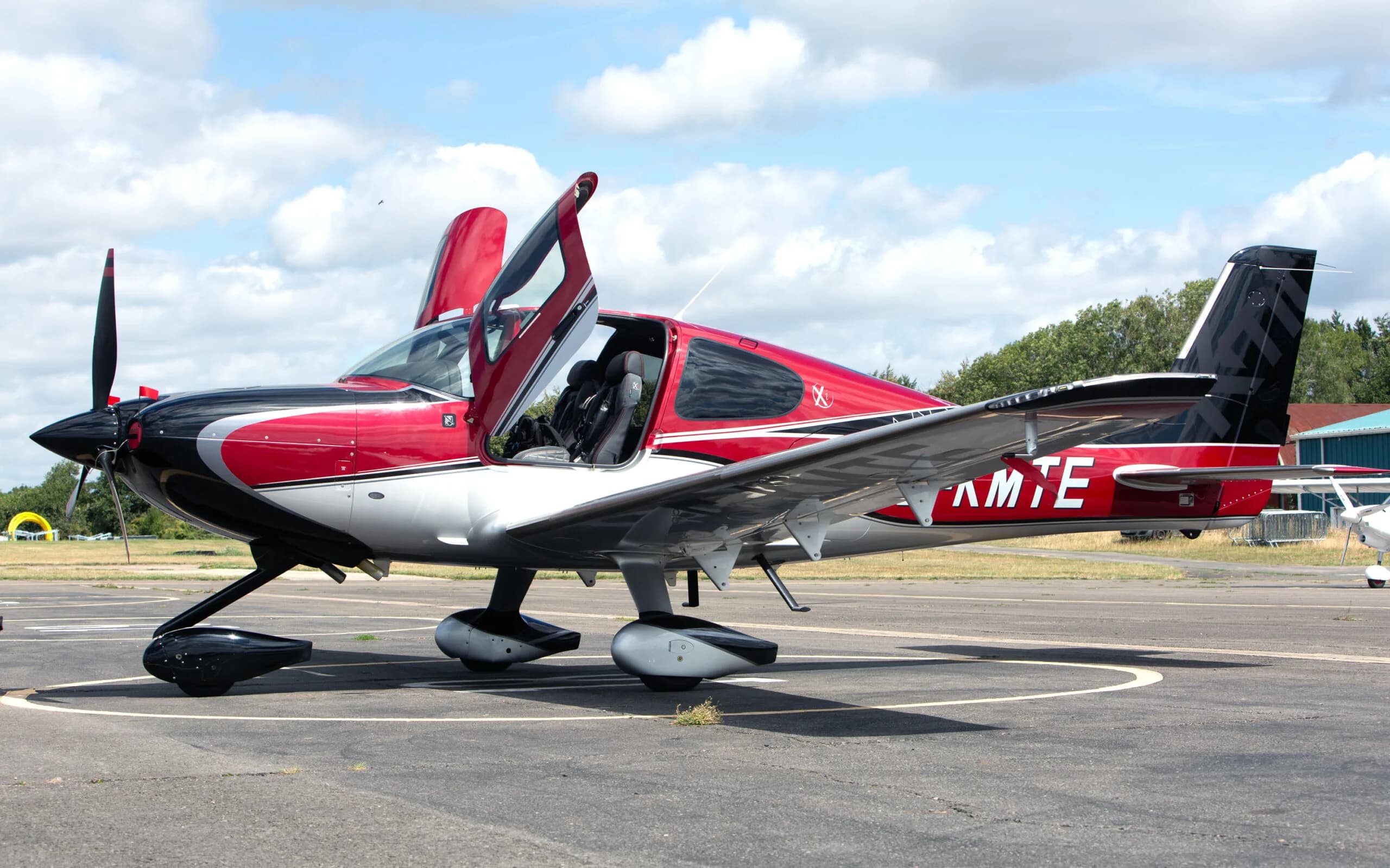 A side view of a red, black, and silver Cirrus SR22 aircraft with its gull-wing doors open, parked on an airfield.