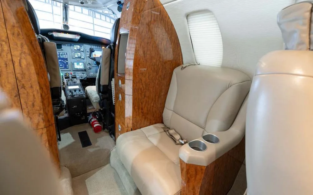 A view from the private jet cabin towards the cockpit, showing a beige seat, wood paneling, and a fire extinguisher.