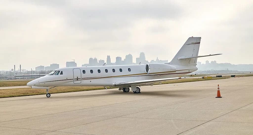 A white business jet with gold and red stripes on the tail sits on an airport tarmac with a city skyline in the background.