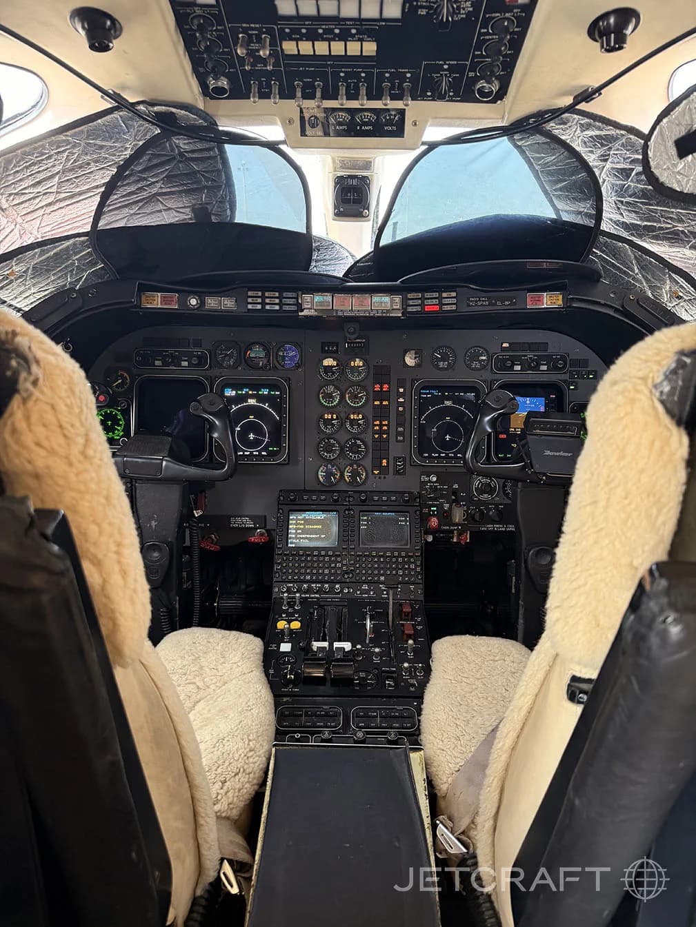 The cockpit of a business jet with multiple screens, instruments, and sheepskin seat covers.