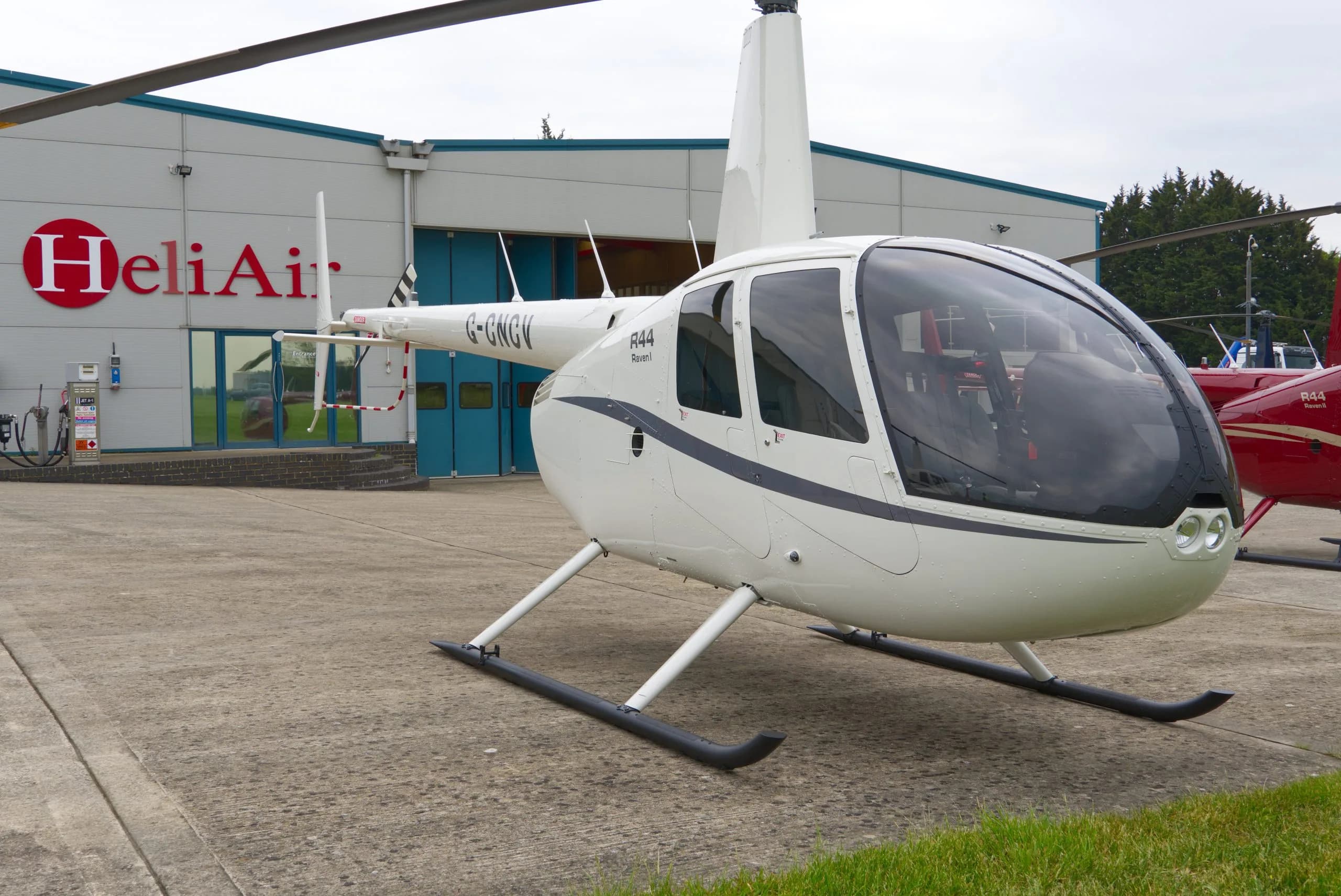 A white Robinson R44 Raven I helicopter (G-GWGV) with dark blue stripes parked on a tarmac in front of a HeliAir hangar.