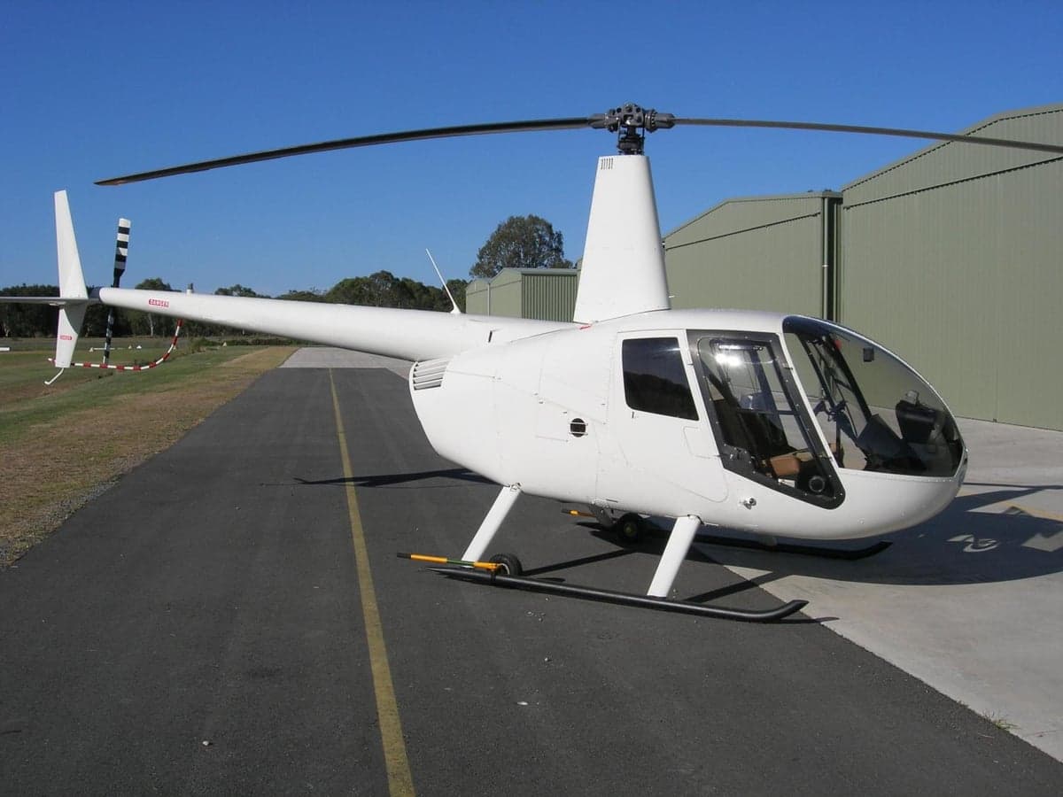 A white Robinson R44 helicopter parked on an asphalt runway next to a green hangar under a clear blue sky.