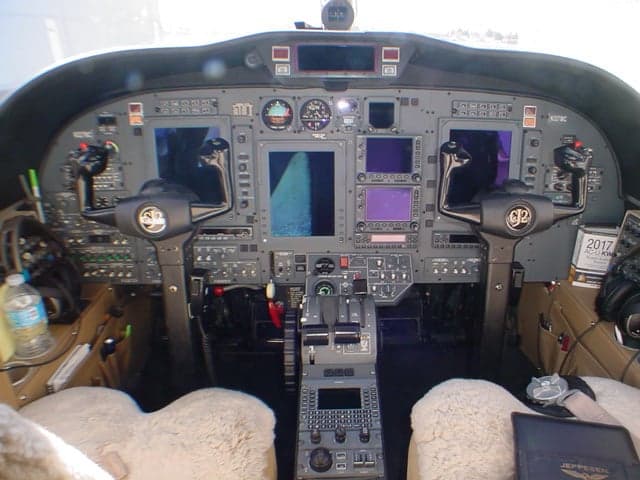 The cockpit of a Cessna Citation jet, showing the instrument panel with multiple screens, controls, and two sheepskin-covered pilot seats.