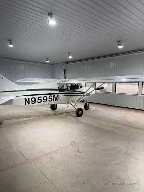 A white Maule M-7-235B aircraft with black stripes and registration N959SM is parked inside a hangar, viewed from the side.