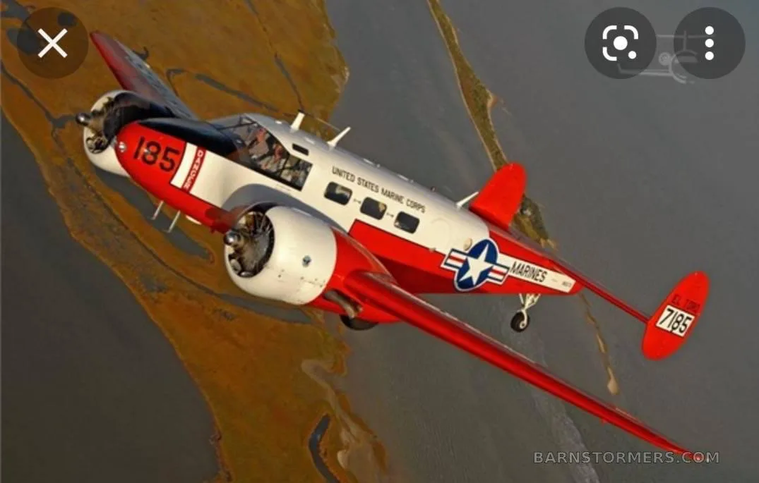 An aerial view of a red and white twin-engine aircraft with 'United States Marine Corps' markings flying over a coastal landscape.