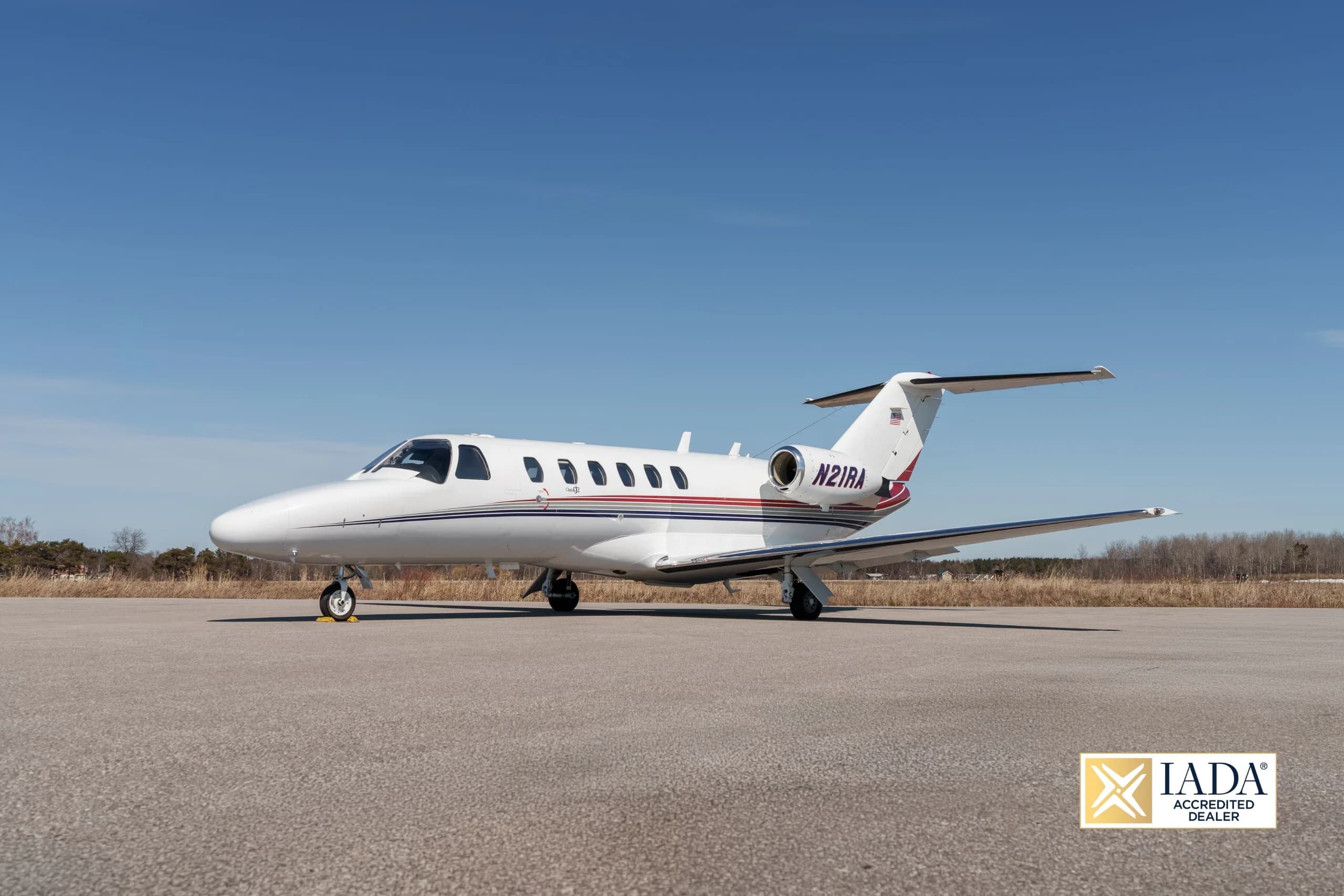 A white Cessna Citation business jet (N21RA) with red and blue stripes parked on an airfield, viewed from the front-left.