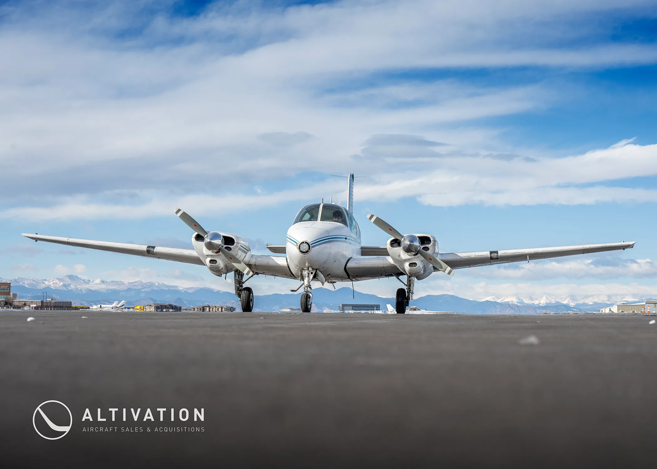 A white and blue twin-engine aircraft is parked on a tarmac with mountains in the background under a cloudy sky.