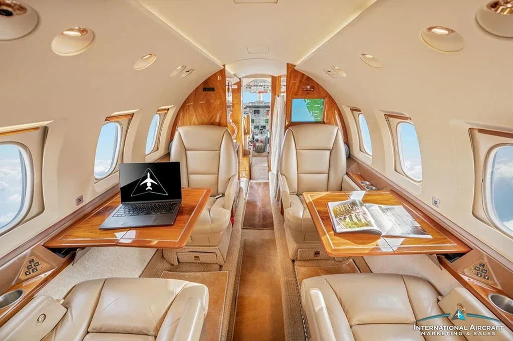 The private jet's cabin features beige leather seating, wooden accents, and a view towards the cockpit.