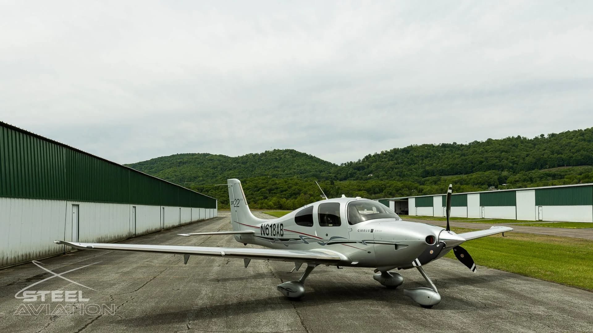 A silver Cirrus SR22 GTS aircraft with registration N618AB parked on an tarmac with hangars and green hills in the background.