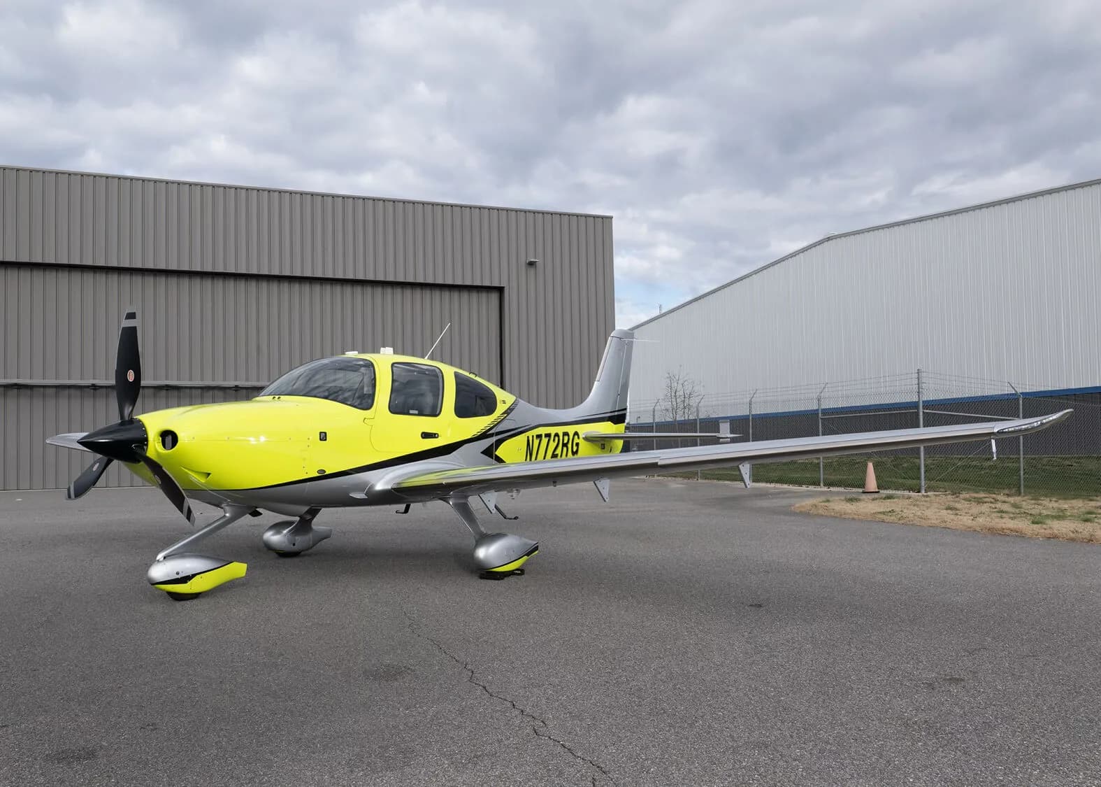 A front three-quarter view of the yellow and silver Cirrus SR22, N772RG, parked on an airfield with hangars in the background.
