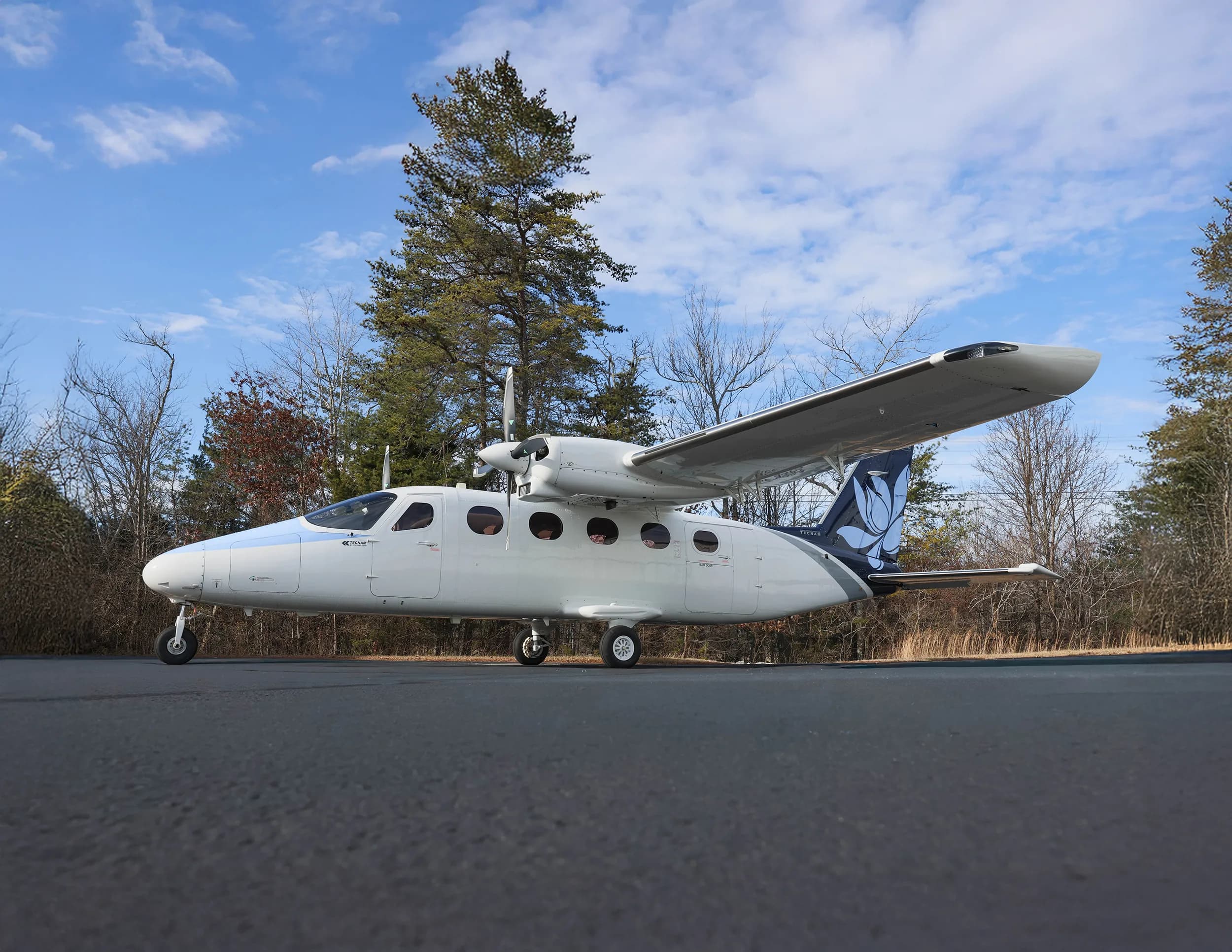 A white and blue twin-engine aircraft with a floral tail logo parked on an asphalt ramp with trees in the background.