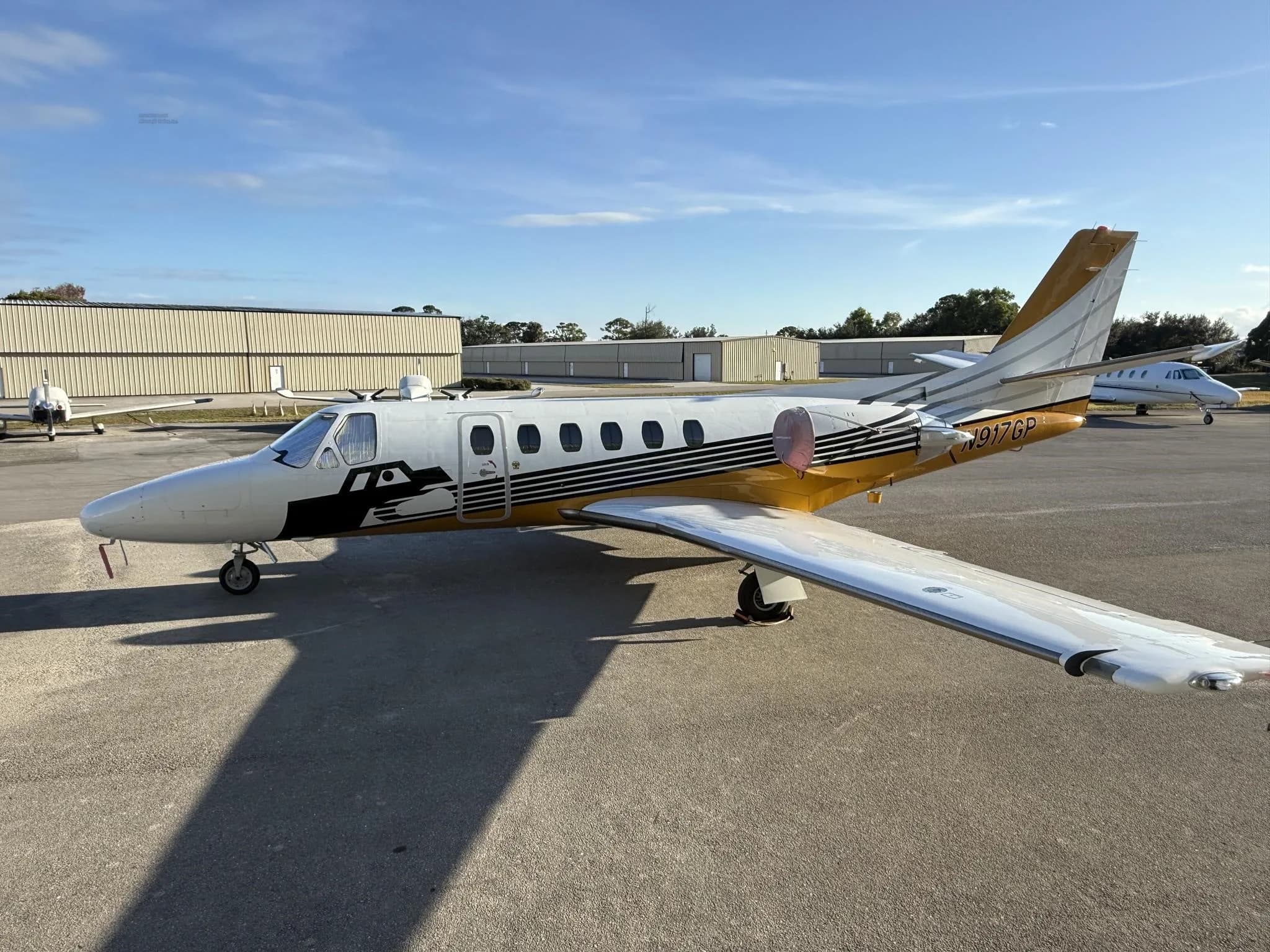 A Cessna Citation jet, N917GP, with white and gold paint scheme, parked on an airfield with other aircraft in the background.