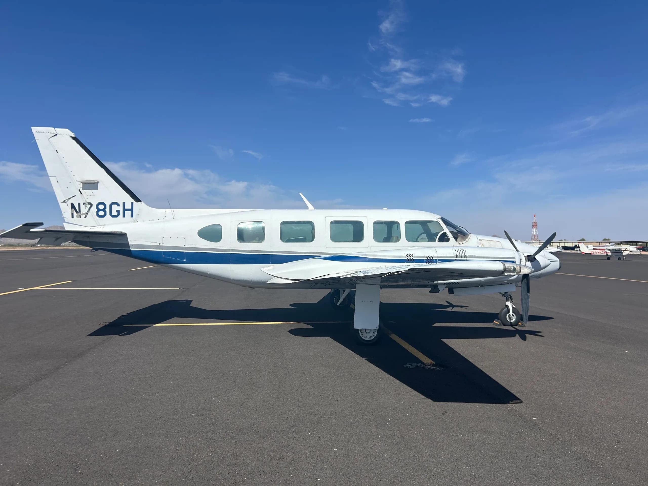 A white twin-engine aircraft with blue and grey stripes, registration N78GH, parked on an asphalt tarmac under a clear blue sky.