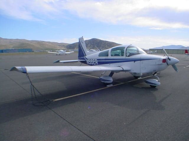 A white and blue Grumman AA-5 Traveler aircraft (N56ER) parked on a tarmac with mountains in the distance.