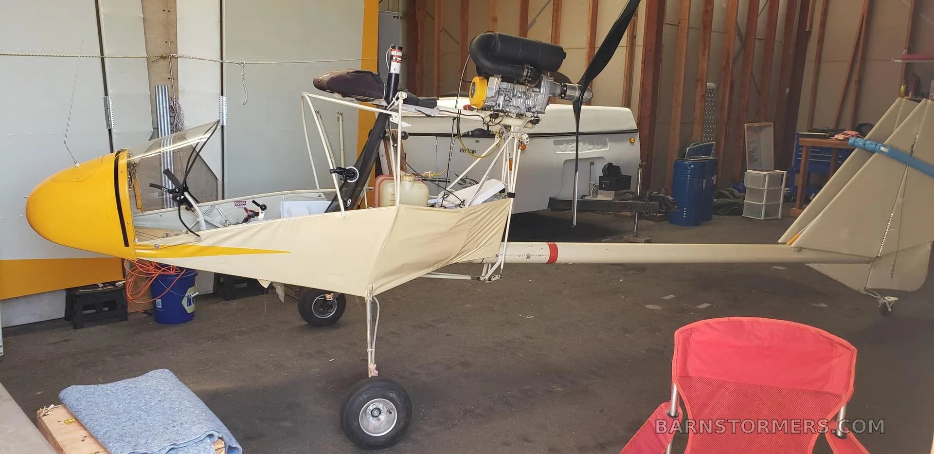A yellow and white ultralight aircraft, partially assembled with its engine exposed, sits in a hangar next to a red chair.